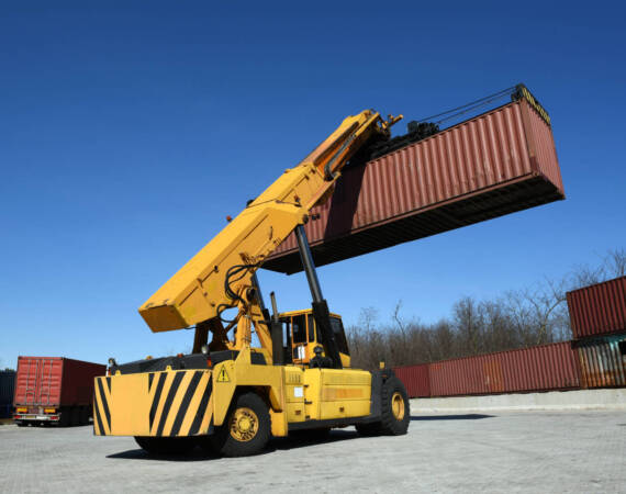 Rear view of yellow container handling vehicle carrying big red container up in the air, low angle view against clear blue sky