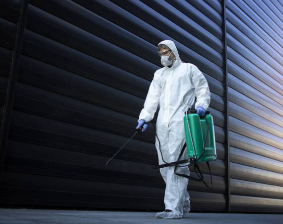 Person in white chemical protection suit doing disinfection of public areas to stop spreading highly contagious corona virus.